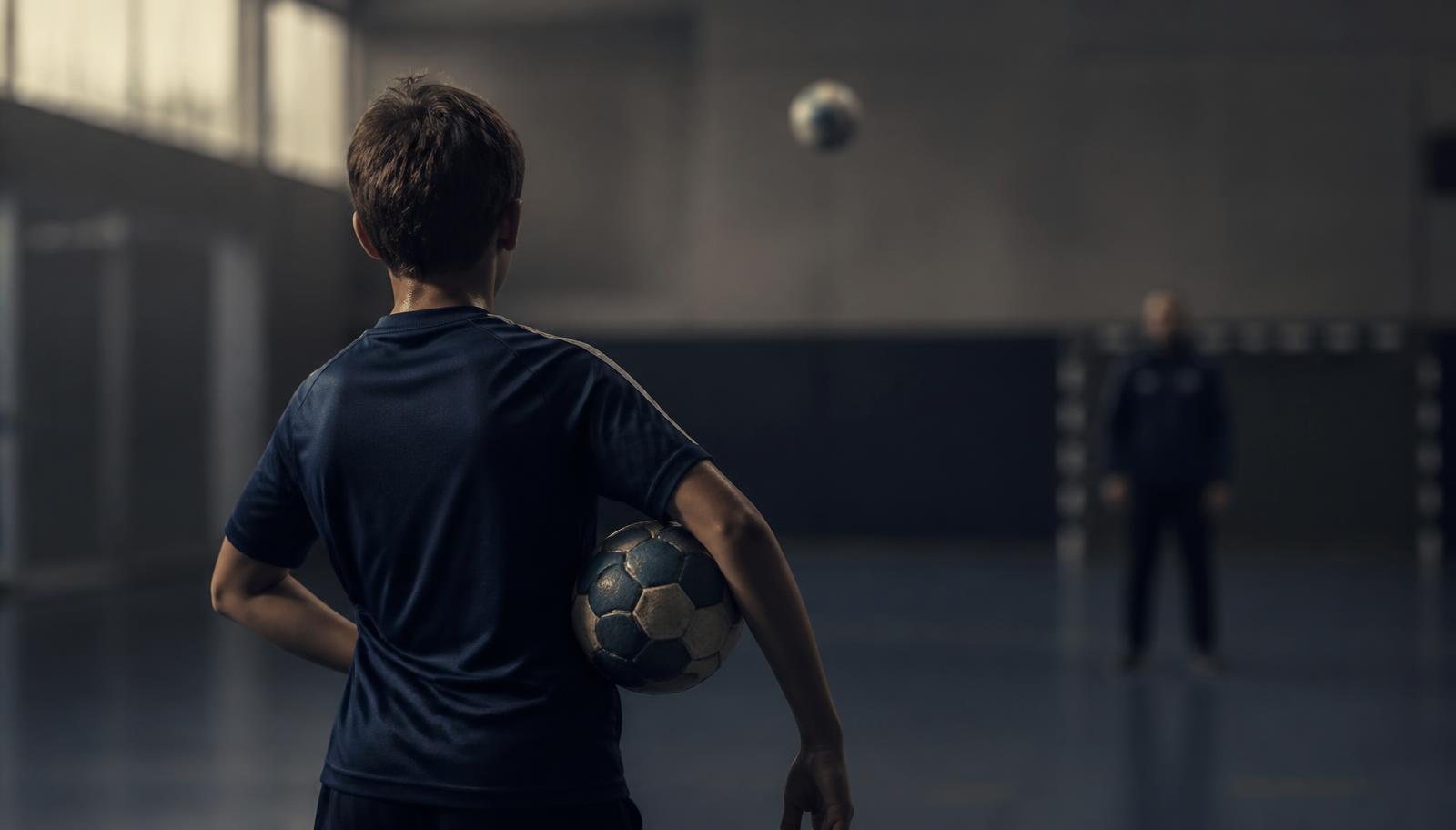 Jovem atleta em quadra observando a trajetória da bola, com o treinador desfocado ao fundo